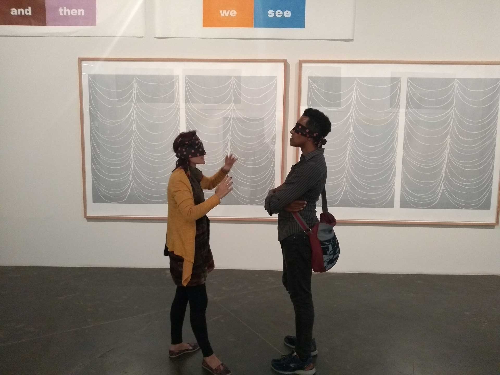 Two people stand blindfolded in front of art exhibition