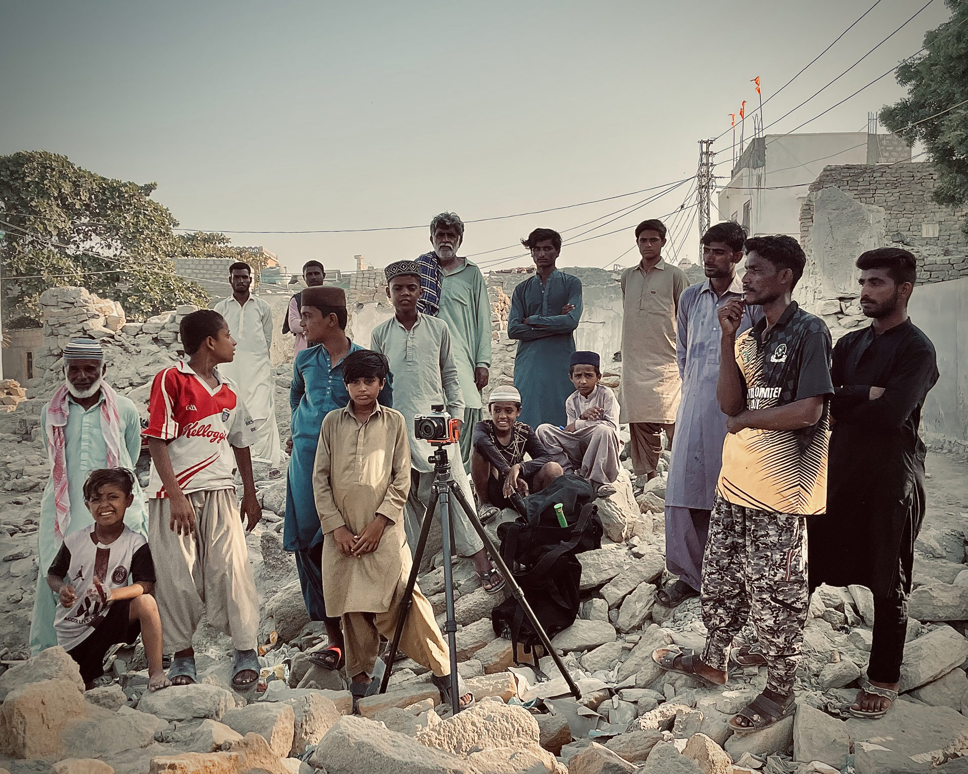 A group of men and boys are standing in what looks like a ruined house, under a blue sky, with a camera on a tripod in front of them. 