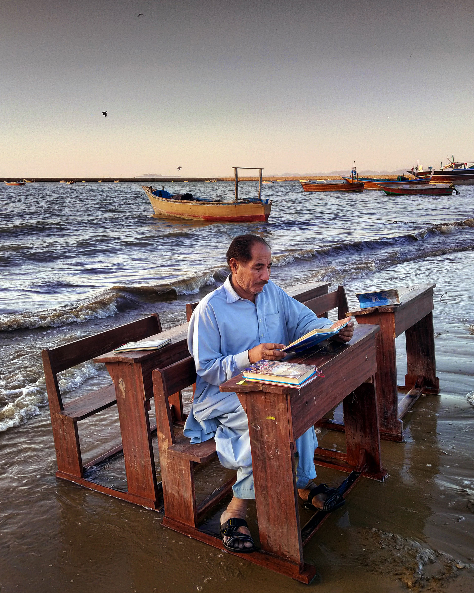A photograph of a man sitting at a wooden table and with an attached bench on a beach. The waves are lapping around him and there are fishing boats moored in the sea behind him. 
