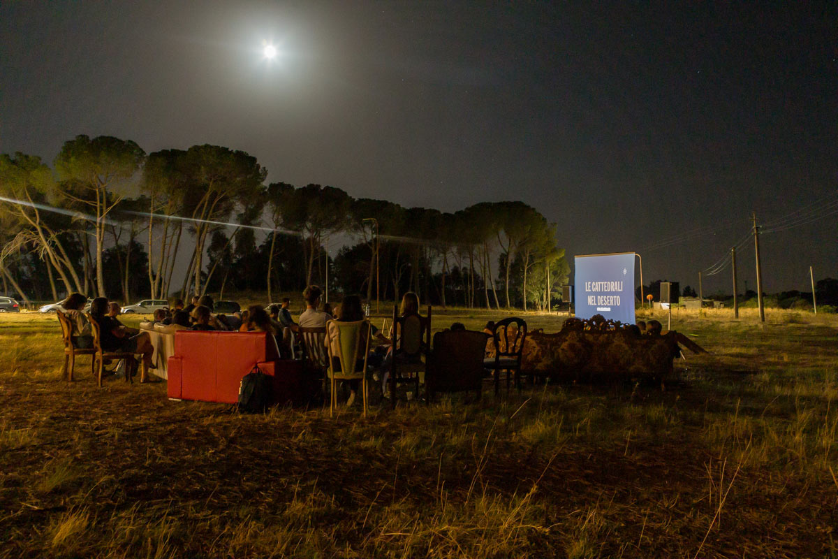 Group of people, sitting in an open field at night watching a film projected on a screen