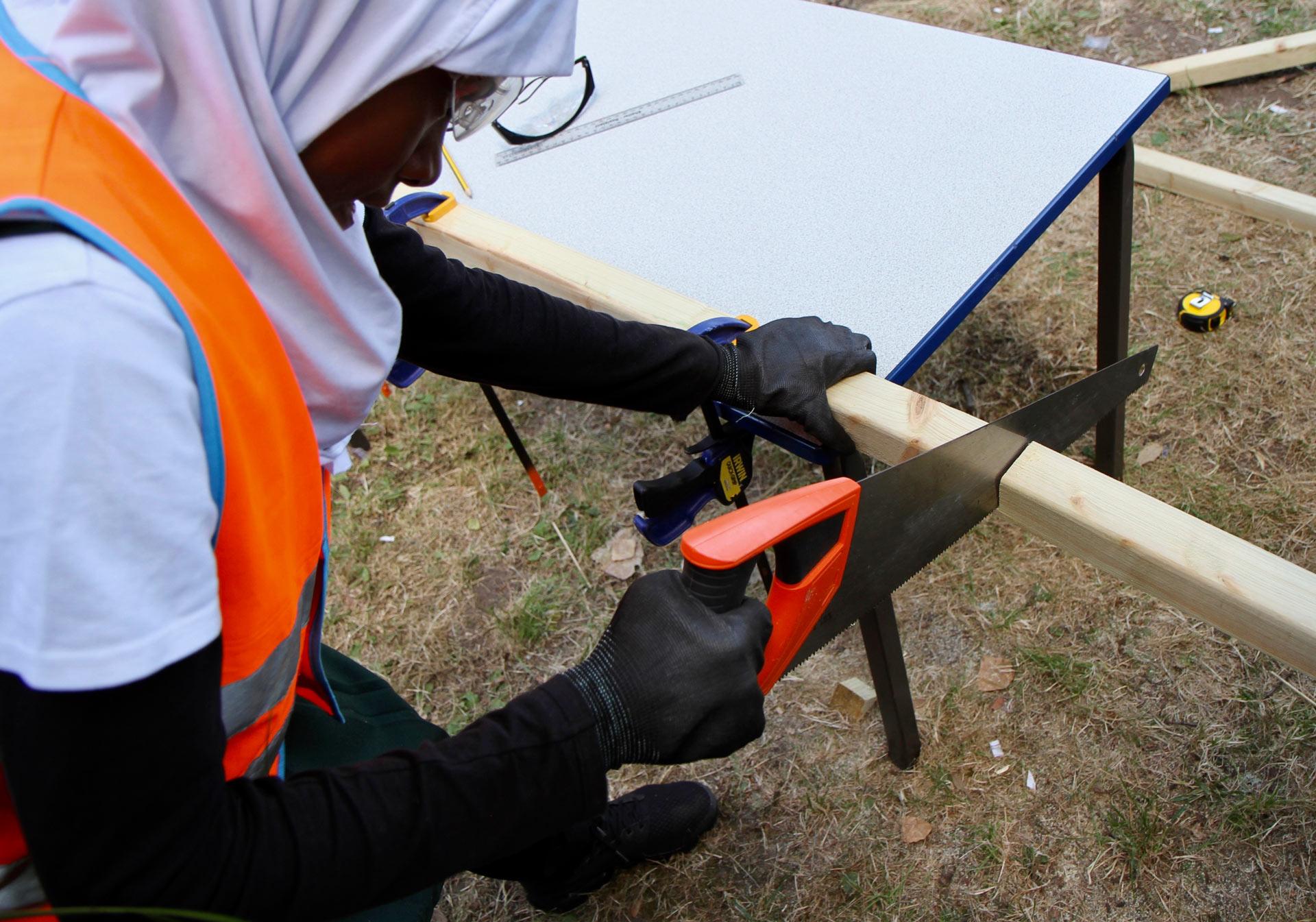 A length of timber is clamped to a classroom table, clutched firmly by a girl wearing a high-vis vest, safety goggles, gloves and a head scarf, who is deftly sawing the timber in two