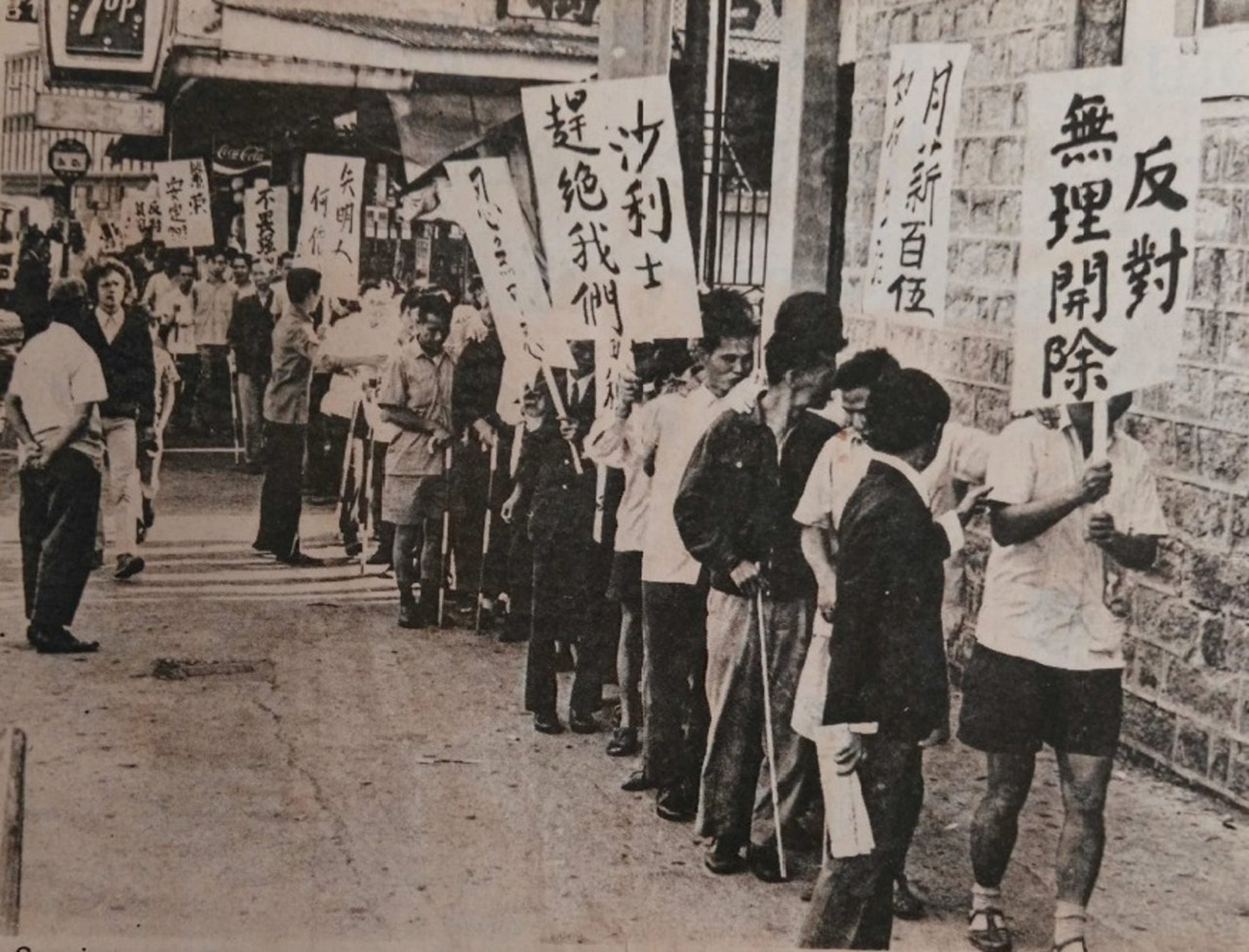 Landscape-orientated news photograph of blind factory workers on strike in 1971. In the photo, from the back left to the front right, there are blind workers lined up for the demonstration. Some of them are holding a cane in one hand and their other hand is on the shoulder of the worker in front of them. Some of them are holding protest signs with various complaints written on them, with Chinese slogans such as “Against Unjustifiable Dismissal”, “Sales Driving Us Out”, “Intolerable”, “What is the Price of Blindness?”, “No Fear of the Strong”, etc. The photo shows the blind workers are walking towards the office of Members of the Executive and Legislative Councils.